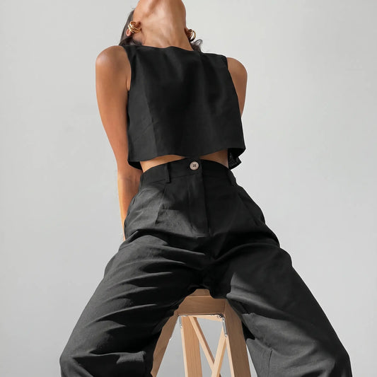 Model wearing a black linen blend cropped sleeveless top and high-waisted wide-leg trousers, styled for effortless summer chic in a studio setting. Features a high neckline and a tailored flat waistband with a single button.