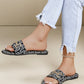 A woman wearing black slide sandals featuring an all-over white logo monogram print and ornate gold-tone hardware, paired with ripped light-wash jeans.
