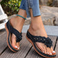 Black Lyraka thong sandals featuring a delicate fabric flower embellishment and gold studs, worn by a model with white nail polish and blue jeans on a wooden deck.