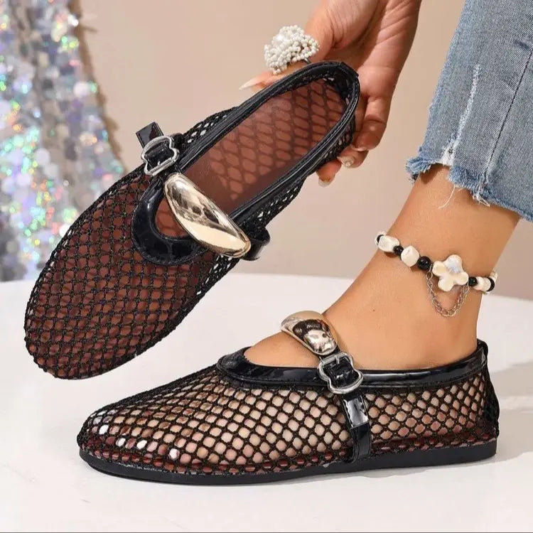 A pair of black mesh flat sandals with patent trim and decorative metallic buckle straps are shown, one worn on a foot with an anklet and the other held by a hand.