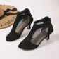 Elegant black suede-like peep-toe ankle boots with sheer mesh side panels and a slim stiletto heel, styled with a straw hat. This three-quarter view showcases the intricate details and chic design.