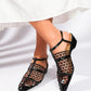 A pair of women's black mesh heeled sandals with pointed toes and thin ankle straps, worn by a model with a white skirt hem, showcasing their elegant design on a studio background.