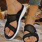 A pair of black mesh wedge sandals with criss-cross straps, worn by a woman with manicured toes and gold anklets, set against a rustic wooden and natural background.