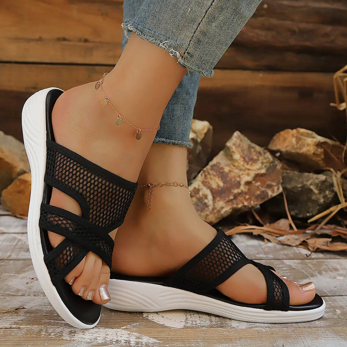 Black mesh wedge sandals on a woman's feet, showcasing the criss-cross straps, contoured white sole, and open-toe design against a rustic wooden and stone background.