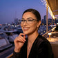 Smiling woman wearing Kivago black metal cat-eye glasses with clear lenses at a waterfront restaurant with city lights.