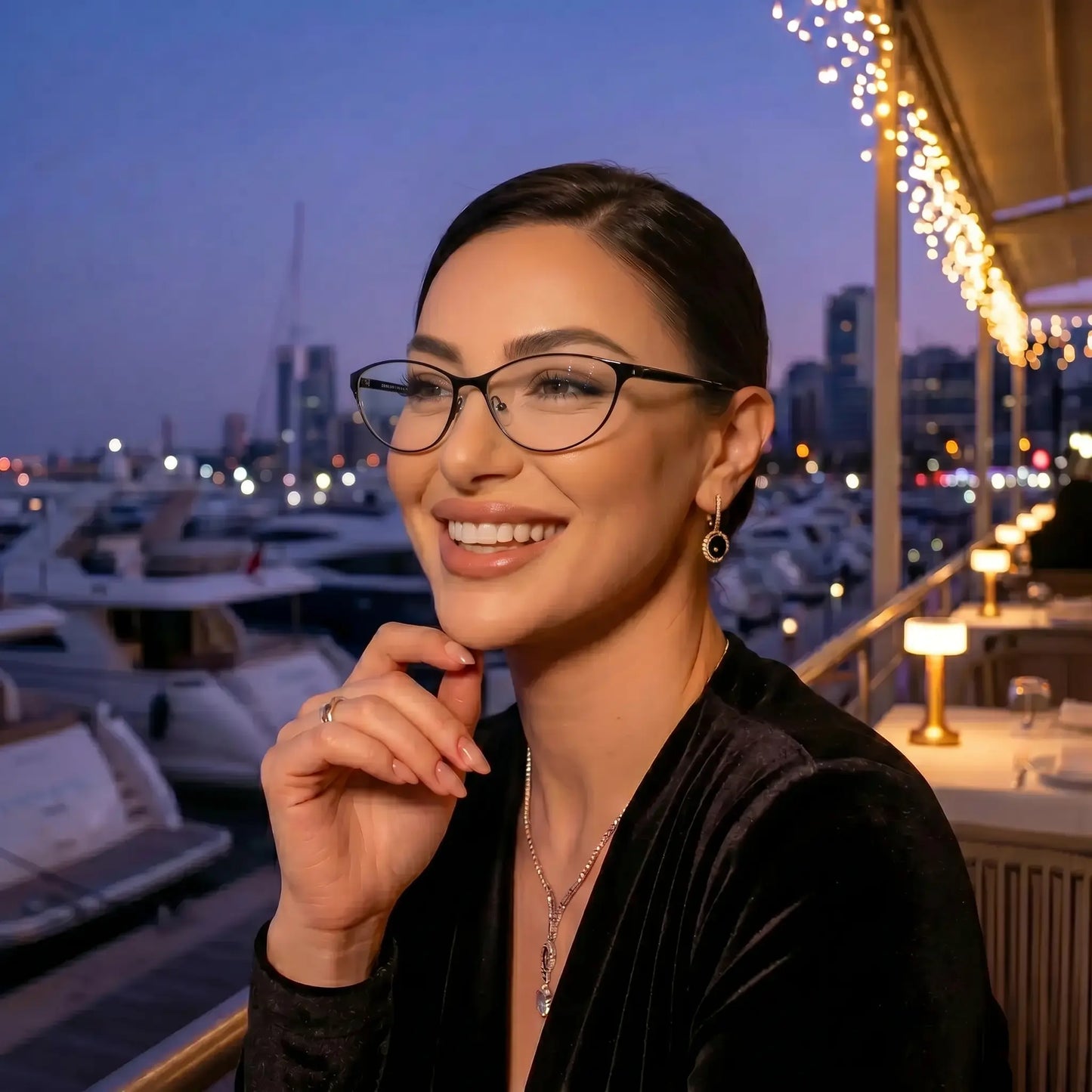 Smiling woman wearing Kivago black metal cat-eye glasses with clear lenses at a waterfront restaurant with city lights.