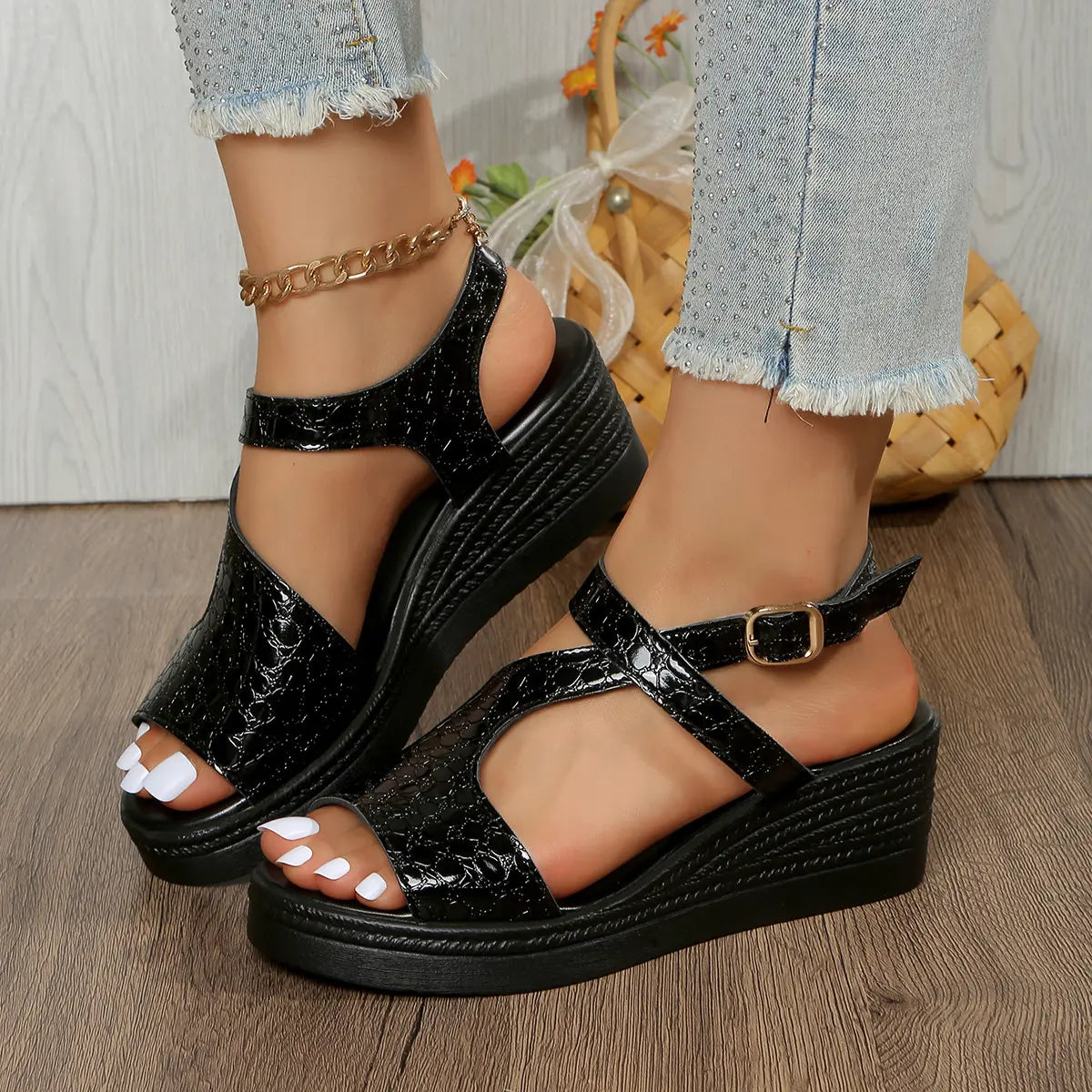 Black metallic peep-toe wedge sandals with an adjustable ankle strap, worn by a model with distressed denim jeans on a wooden floor.