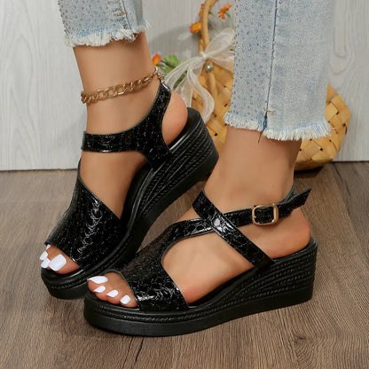 Black metallic peep-toe wedge sandals with an adjustable ankle strap, worn by a model with distressed denim jeans on a wooden floor.