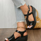 Black metallic peep-toe wedge sandals with a textured finish, worn by a model with distressed denim jeans and a gold ankle bracelet on a wooden floor.