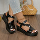 Black metallic peep-toe wedge sandals with an embossed crocodile texture, T-strap, and adjustable ankle strap worn by a model with distressed denim jeans on a wooden floor.