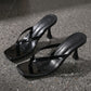 A pair of black patent leather heeled flip-flop sandals with square toes and sculptural stiletto heels, shown from a three-quarter angle on a speckled grey studio surface.