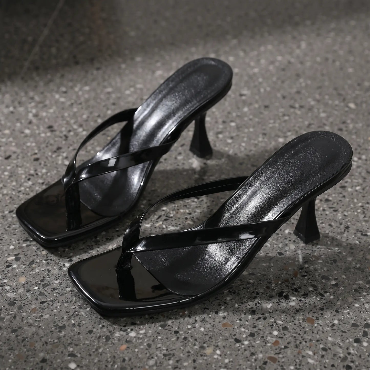 A pair of black patent leather heeled flip-flop sandals with square toes and sculptural stiletto heels, shown from a three-quarter angle on a speckled grey studio surface.