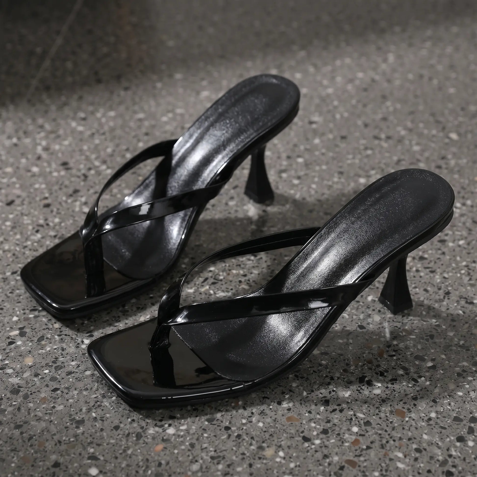 A pair of black patent leather heeled flip-flop sandals with square toes and sculptural stiletto heels, shown from a three-quarter angle on a speckled grey studio surface.