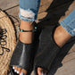 Black perforated PU leather women's flat peep-toe sandals with a single wide strap, worn with distressed jeans on a wooden deck.