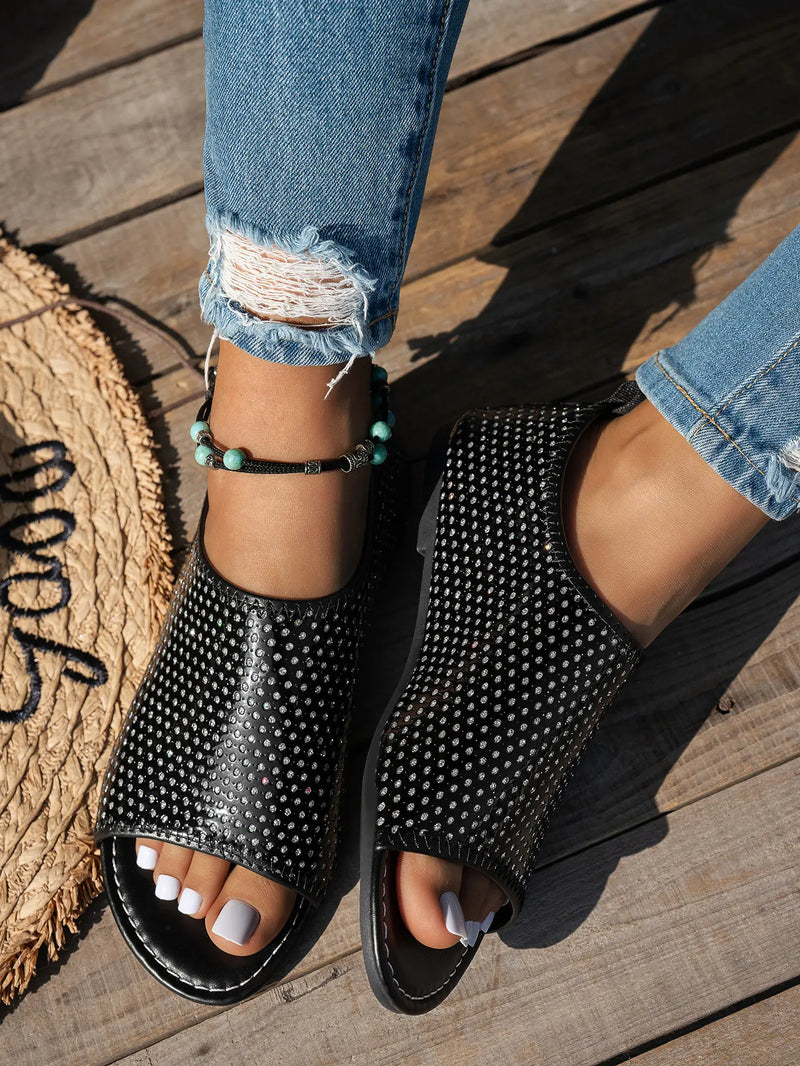 Black perforated PU leather women's flat peep-toe sandals with a single wide strap, worn with distressed jeans on a wooden deck.