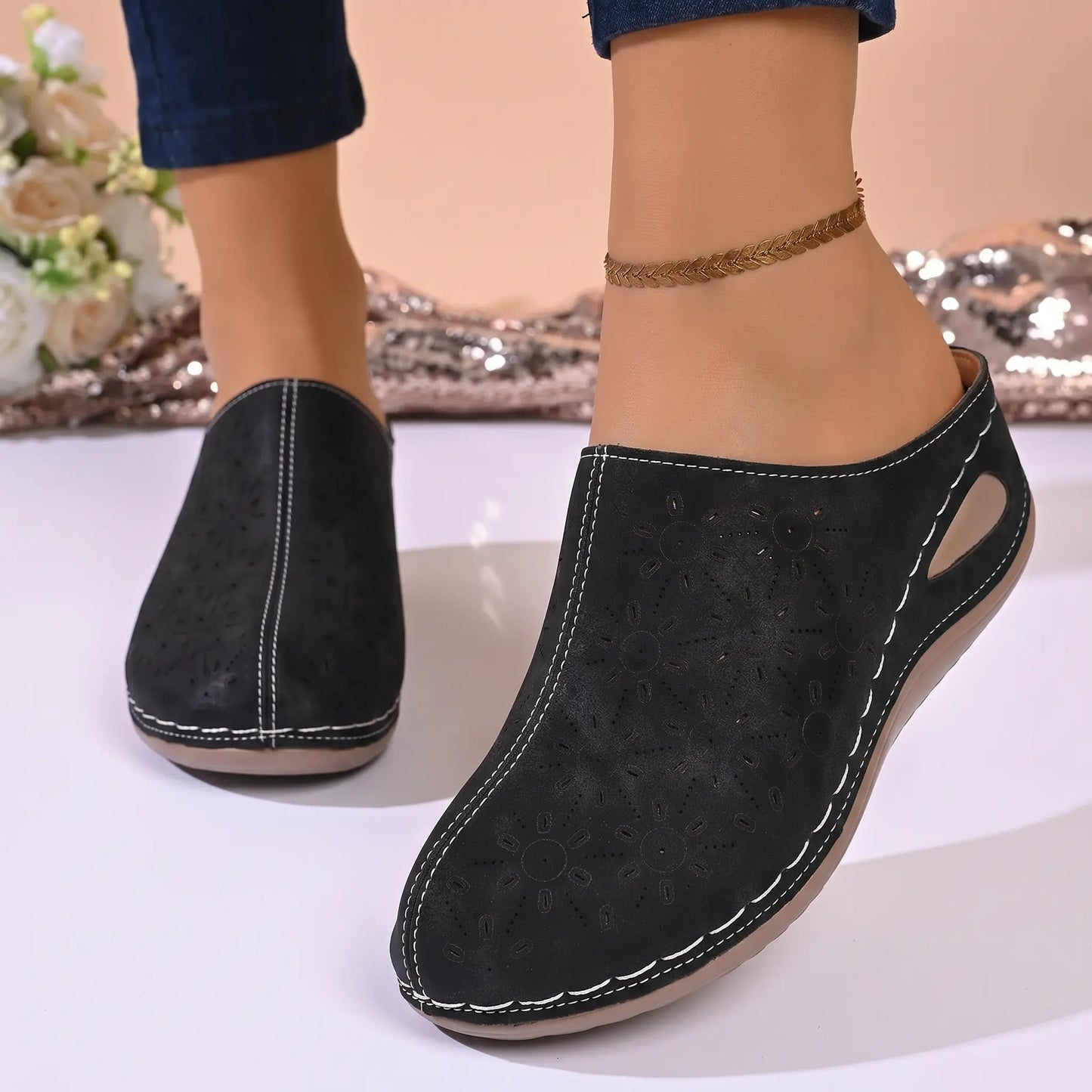 A pair of black Vylos perforated floral wedge mules worn by a model, showcasing the slip-on design, round toe, and white stitching details.