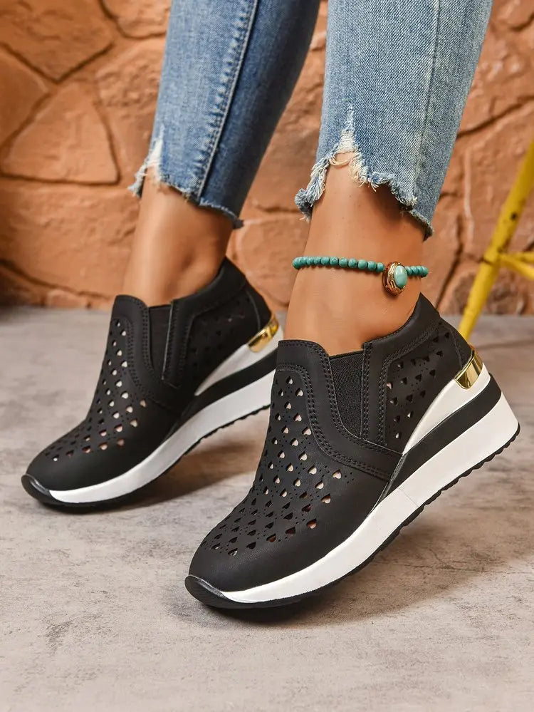 Women's black perforated wedge loafers with a subtle gold heel accent, styled with ripped blue jeans and a turquoise anklet. Features heart-shaped cutouts for breathability and comfort.