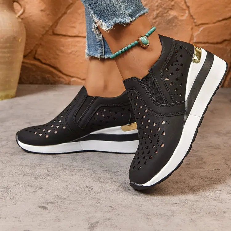 Side view of black perforated heart slip-on wedge loafers worn by a model with ripped jeans and a turquoise anklet.