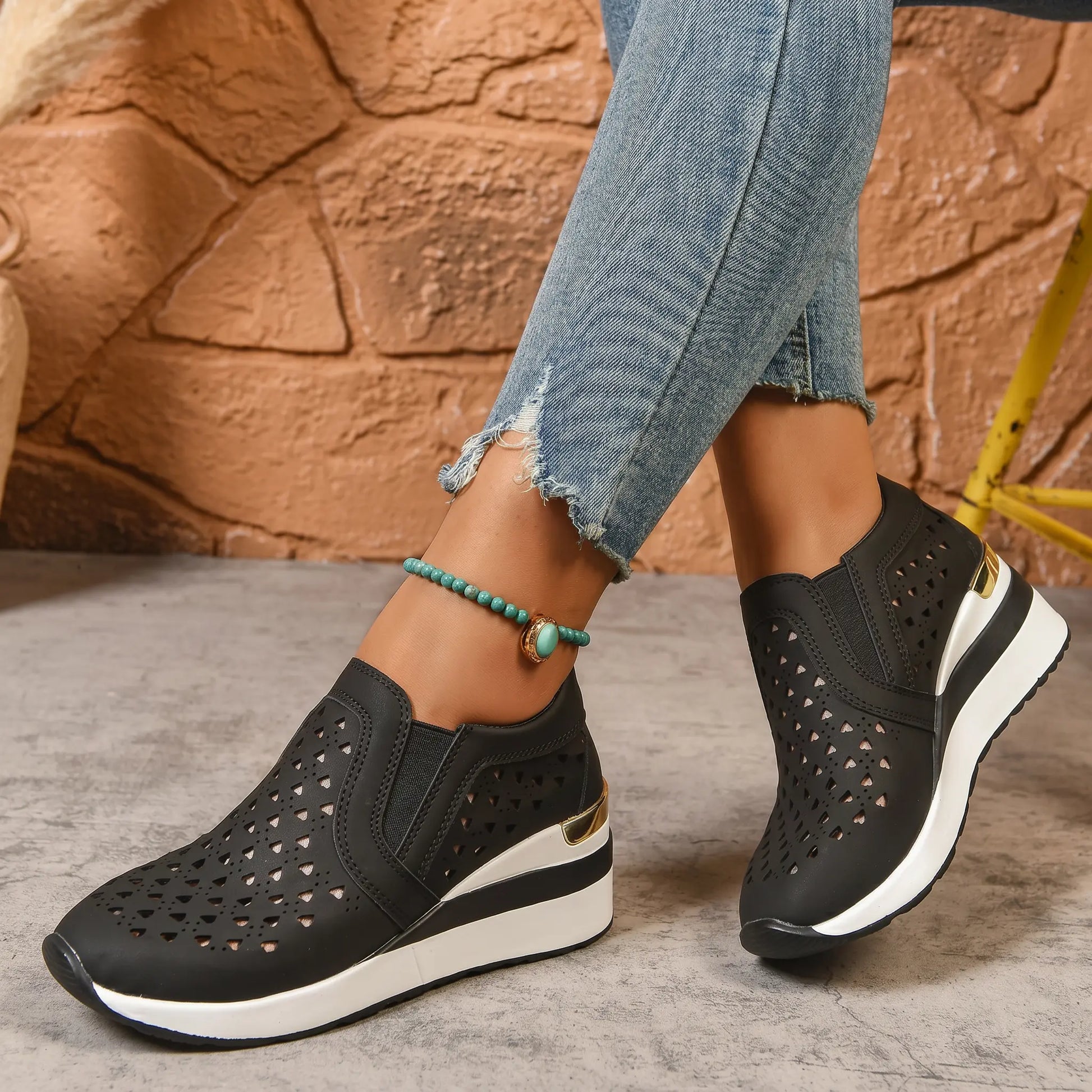 Black perforated heart slip-on loafers with a hidden wedge heel and gold accent, worn with distressed denim jeans and a turquoise anklet.