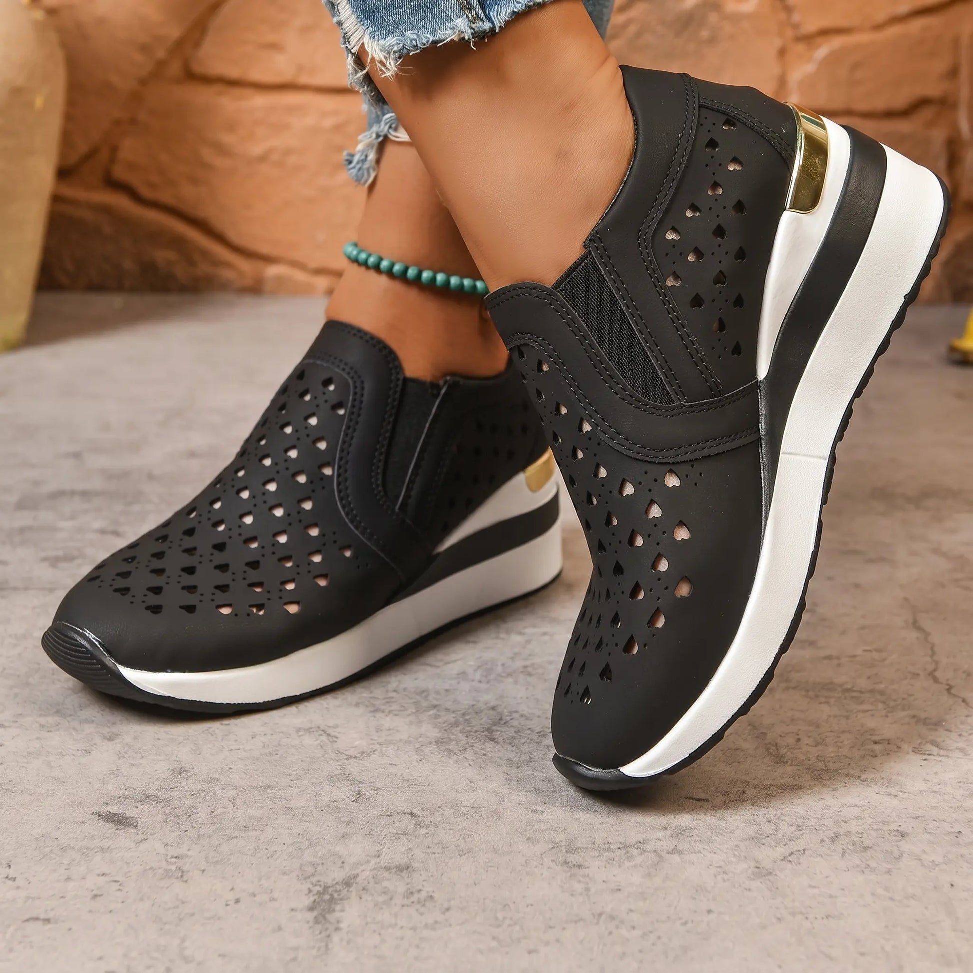 Black perforated slip-on wedge loafers with a subtle gold heel accent, shown on a woman's feet with ripped jeans and a turquoise anklet.