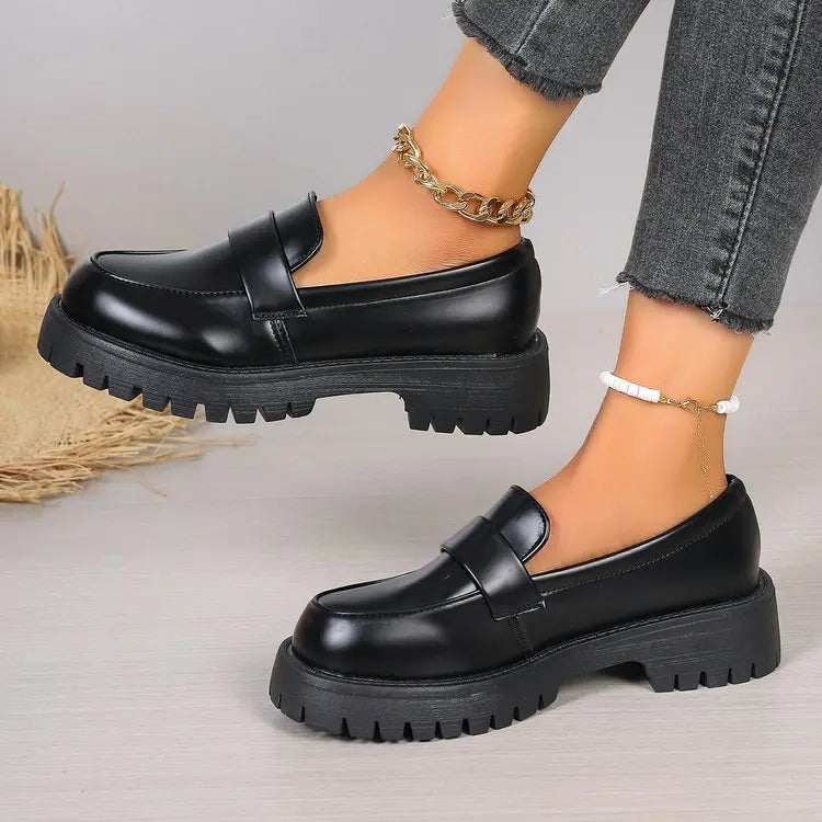 Black polished synthetic leather platform loafers with a chunky lug sole and classic penny strap, shown on a woman's feet with denim jeans.