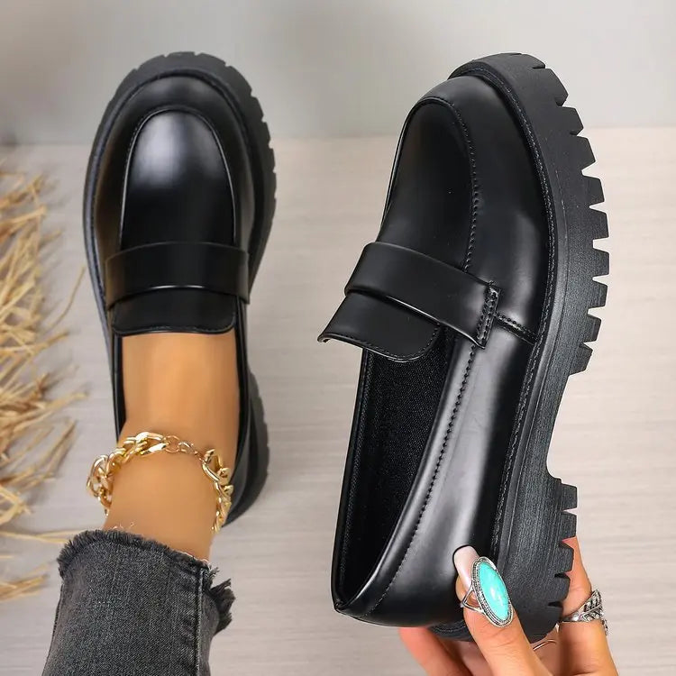 Black polished synthetic leather platform loafers with a chunky lug sole and classic penny strap, shown on a person's feet with a gold ankle bracelet. The background is a light wood surface with blurred natural reeds.