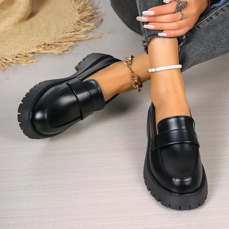 Black polished synthetic leather platform loafers with a chunky lug sole, worn by a model showing the classic penny strap detail and round toe. Gold chain and white beaded anklets add styling.