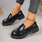Women's black polished synthetic leather platform loafers with a chunky lug sole and classic penny strap, shown on feet with decorative anklets.