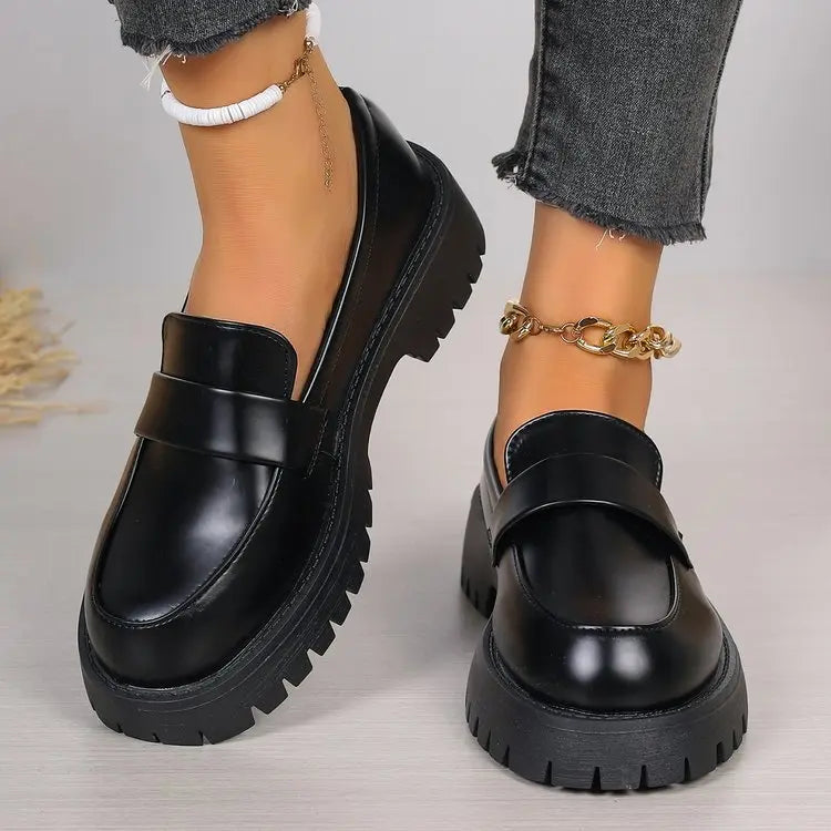 Black polished synthetic leather platform loafers for women, featuring a chunky lug sole and classic penny strap detail. Presented in a three-quarter view on a neutral studio background.