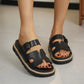 Black vegan leather platform slides featuring an adjustable buckle, distinctive H-cutout straps, and a layered sole with tan cork detail and white contrast stitching, worn by a model.