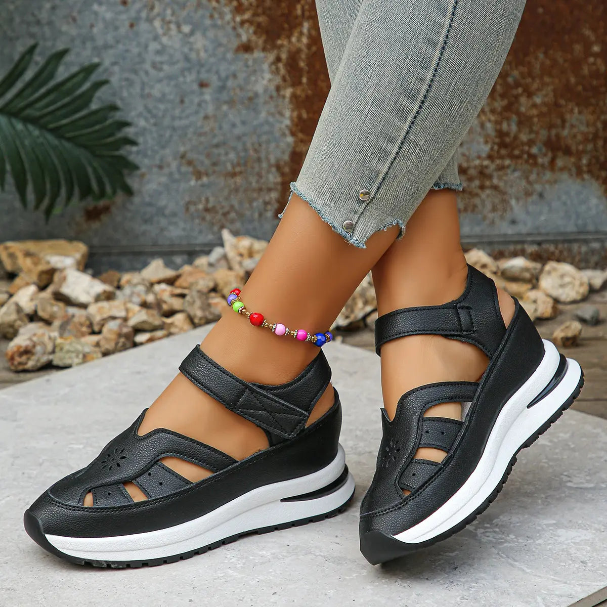Black platform wedge sandals with cut-out straps and a white midsole, worn by a woman with jeans and a colorful anklet in a lifestyle setting.