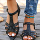 Elegant black flat sandals with a pointed toe, delicate T-strap, and elasticated slingback strap, shown on a model's feet wearing distressed denim jeans on a wooden deck.