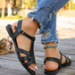 A woman's feet wearing black pointed-toe flat sandals with an elasticated slingback strap and T-strap design, styled with distressed jeans on a wooden deck.