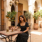 Smiling woman in a black polka dot maxi dress featuring a square neck and puff sleeves, seated at an outdoor cafe table in a sunlit courtyard.
