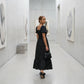 Woman wearing a black Buzalo polka dot smocked maxi dress with puff sleeves and a tiered A-line skirt, glancing back in a modern art gallery.