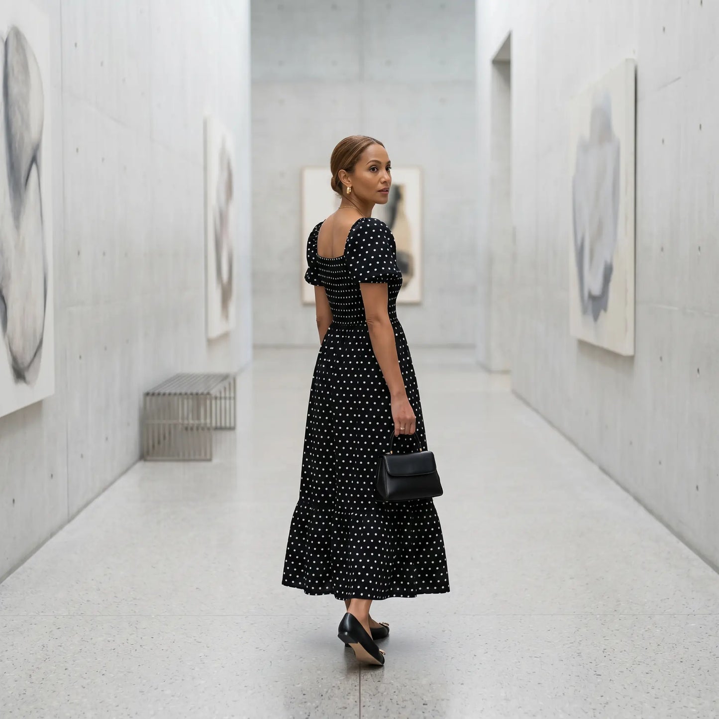Woman wearing a black Buzalo polka dot smocked maxi dress with puff sleeves and a tiered A-line skirt, glancing back in a modern art gallery.