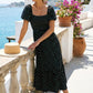 Elegant model wearing a black polka dot maxi dress featuring a smocked square neckline, puff sleeves, and a tiered hem, posing on a sunlit Mediterranean balcony overlooking the sea.