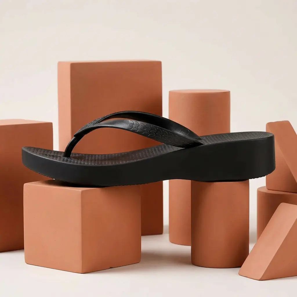 Black waterproof PVC platform wedge flip-flop, presented in a side three-quarter view, showcasing its embossed leaf straps and honeycomb textured footbed on studio props.