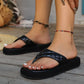 A woman's feet wearing black quilted PU platform thong sandals with white toenails and colorful beaded anklets, set against a rustic wooden floor background.