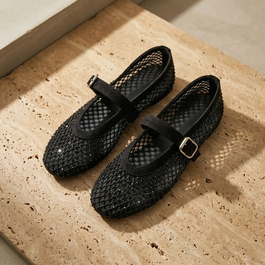 A pair of Galdyr Midnight Black Rhinestone Mesh Mary Jane flats with rounded toes, suede-like trim, and a silver buckle strap, viewed from a three-quarter angle on a textured beige surface.