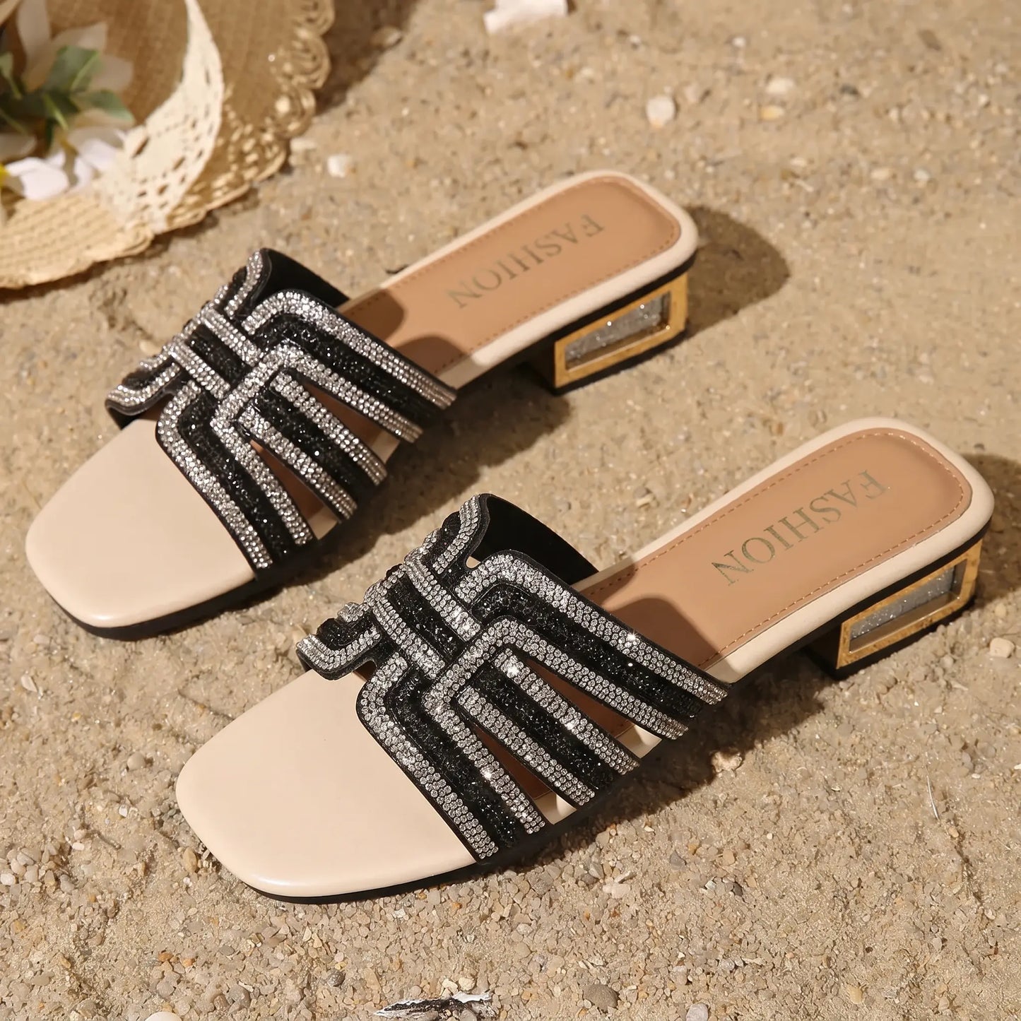 Black embellished slide sandals with square toes and intricate geometric rhinestone straps, resting on a sandy beach next to a straw hat, showcasing luxury summer footwear.