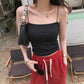 Woman wears a black slim-fit camisole with thin spaghetti straps, a straight neckline, and subtle ribbed texture. She holds black sunglasses, showcasing the top styled with red drawstring pants.