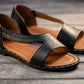 Close-up view of black faux leather Roman flat sandals featuring a geometric cross-strap design, tan footbed, and decorative silver-tone studs along the sole edge, displayed on a rustic wooden surface.
