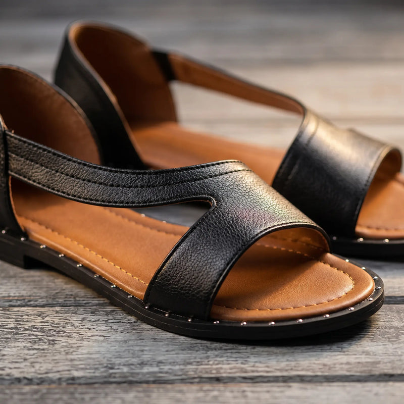 Close-up view of black faux leather Roman flat sandals featuring a geometric cross-strap design, tan footbed, and decorative silver-tone studs along the sole edge, displayed on a rustic wooden surface.