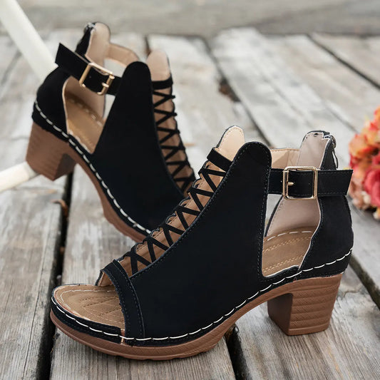 Black faux suede Roman sandals with criss-cross lace-up front, open toe, chunky block heel, and gold buckle ankle strap, set on a rustic wooden surface.