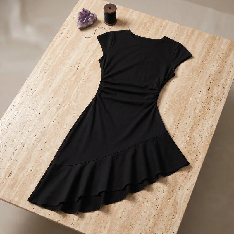 Black Jaryth Premium Cotton Ruched Asymmetrical Dress, flat-laid on a light stone surface, highlighting its cap sleeves, ruched waist, and ruffled asymmetrical hem.
