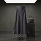 Elegant black satin high-waisted midi skirt with an A-line mermaid drape, styled in a dark studio setting with a coordinating handbag and shoes.