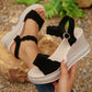 Women's black suede-like wedge sandals with scalloped straps and rhinestone-trimmed platform, worn on feet in a natural outdoor setting.