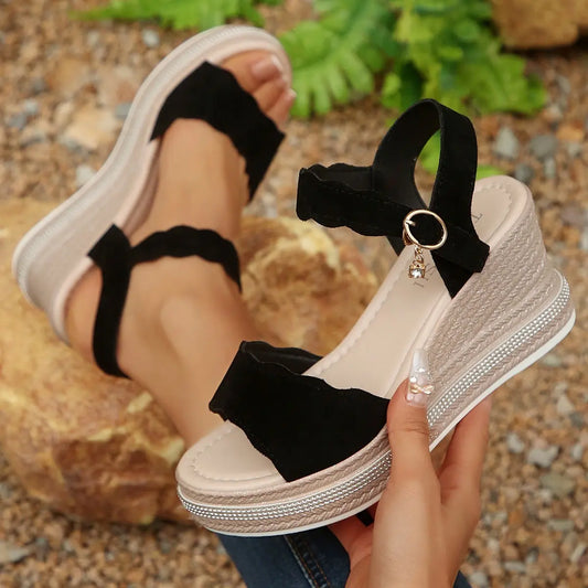 Women's black suede-like wedge sandals with scalloped straps and rhinestone-trimmed platform, worn on feet in a natural outdoor setting.