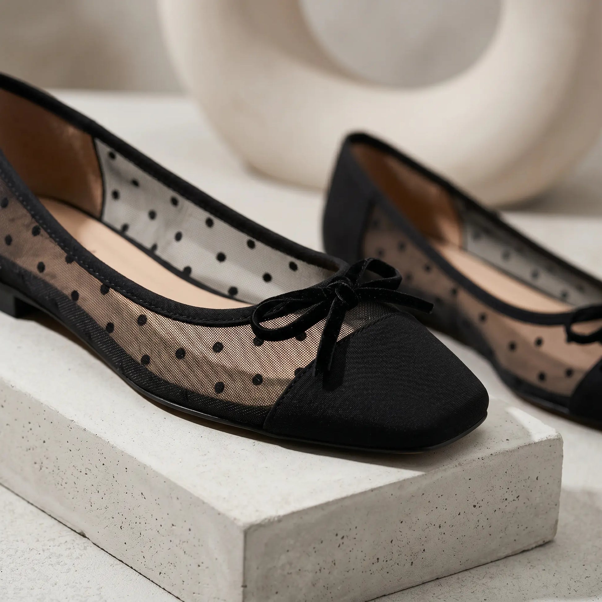 Close-up of black sheer mesh polka dot ballet flats, highlighting the solid square toe cap and delicate velvet bow accent on a studio surface.